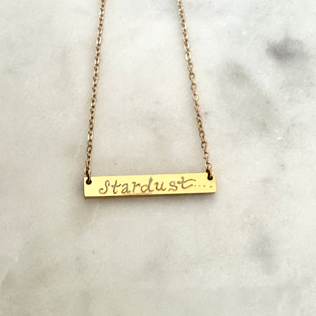 Gold bar necklace with 'Stardust' script on a marble surface